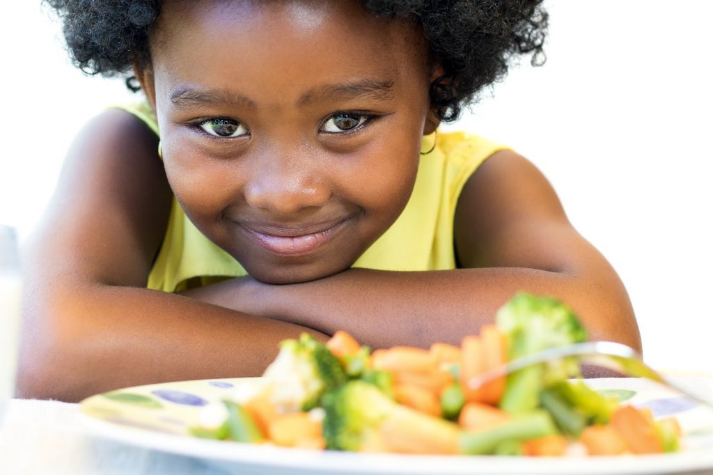 child smiling next to plate of cooked veggies